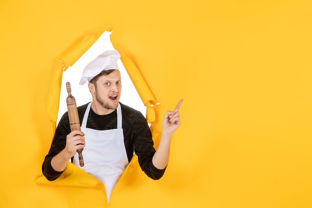 Vista frontal joven cocinero en capa blanca sosteniendo un rodillo sobre el fondo amarillo comida hombre blanco foto color trabajo de cocina