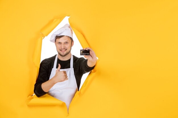 Vista frontal joven cocinero en capa blanca sosteniendo una tarjeta bancaria negra sobre un fondo amarillo cocina de color blanco trabajo hombre comida cocina