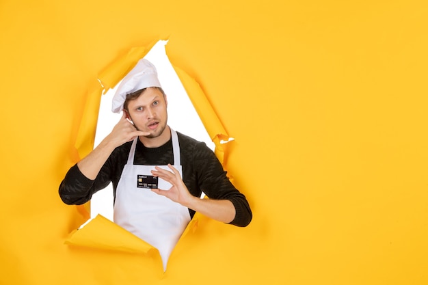 Vista frontal joven cocinero en capa blanca con tarjeta bancaria negra sobre fondo amarillo modelo cocina color blanco trabajo hombre comida dinero