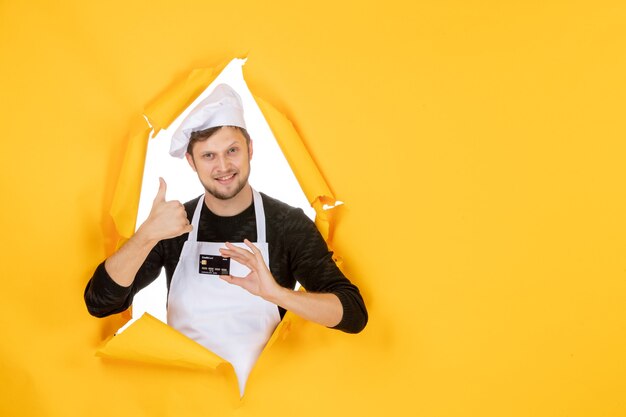 Vista frontal joven cocinero en capa blanca con tarjeta bancaria negra sobre fondo amarillo modelo color blanco trabajo hombre comida dinero