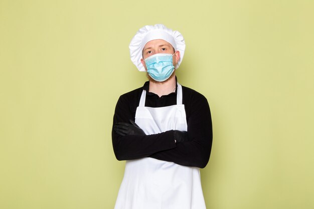 Una vista frontal joven cocinero masculino en camisa negra con capa blanca gorra blanca en guantes negros máscara protectora azul