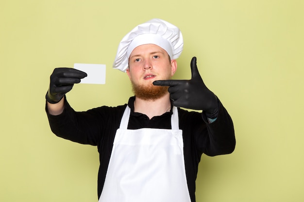 Una vista frontal joven cocinero masculino en camisa negra con capa blanca gorra blanca en guantes negros con tarjeta gris