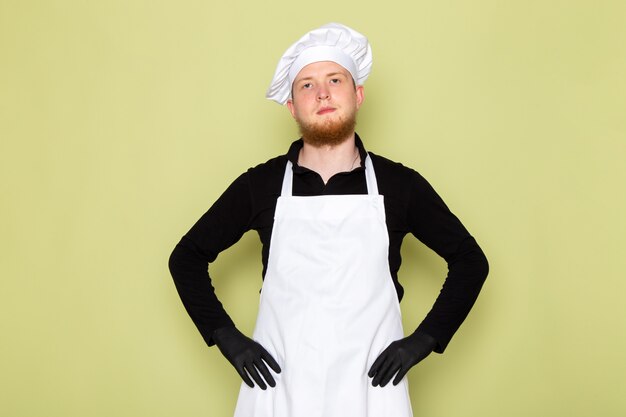 Una vista frontal joven cocinero masculino en camisa negra con gorra blanca cabeza blanca en guantes negros posando