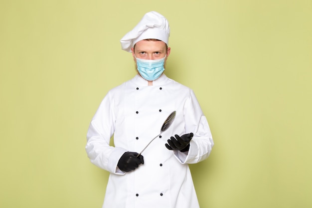 Foto gratuita una vista frontal joven cocinero masculino en traje de cocinero blanco gorra blanca en guantes negros máscara protectora azul con gran cuchara metálica
