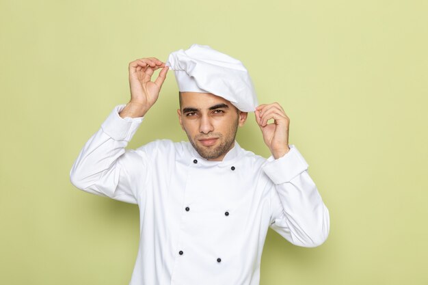 Vista frontal joven cocinero en traje de cocinero blanco arreglando su gorra blanca en verde