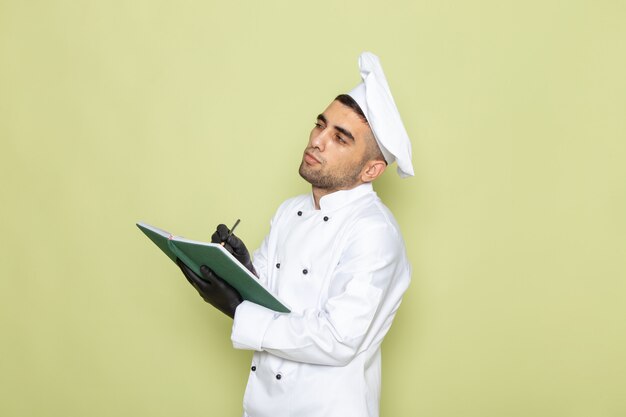 Vista frontal joven cocinero en traje de cocinero blanco con guantes oscuros y anotando notas en verde