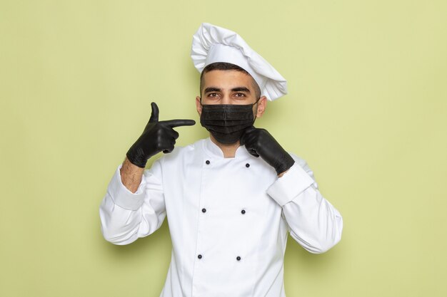 Vista frontal joven cocinero en traje de cocinero blanco con guantes oscuros y máscara estéril en verde