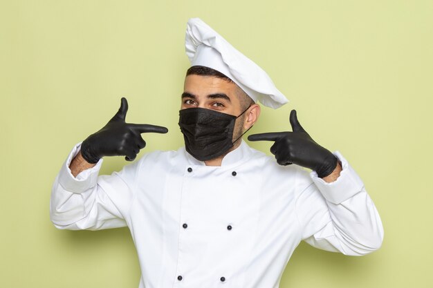 Vista frontal joven cocinero en traje de cocinero blanco con máscara y guantes en verde