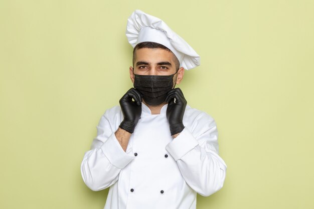 Vista frontal joven cocinero en traje de cocinero blanco con máscara y guantes en verde