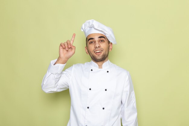 Vista frontal joven cocinero en traje de cocinero blanco posando con el dedo levantado en verde