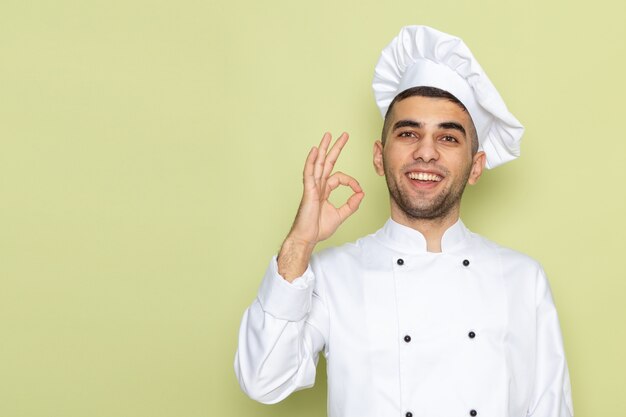 Vista frontal joven cocinero en traje de cocinero blanco sonriendo y mostrando bien firmar en verde