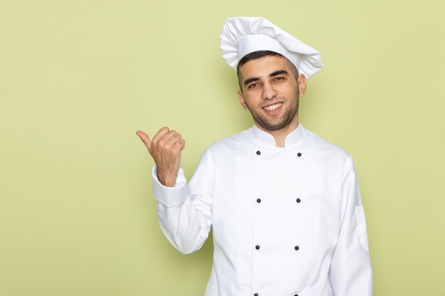 Vista frontal joven cocinero en traje de cocinero blanco sonriendo y mostrando el signo del dedo en verde