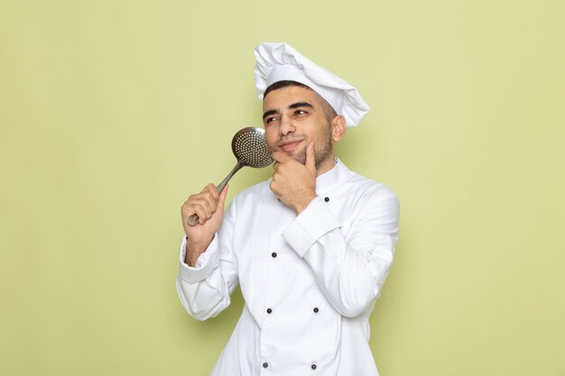 Vista frontal joven cocinero en traje de cocinero blanco sosteniendo una cuchara de plata y pensando en verde