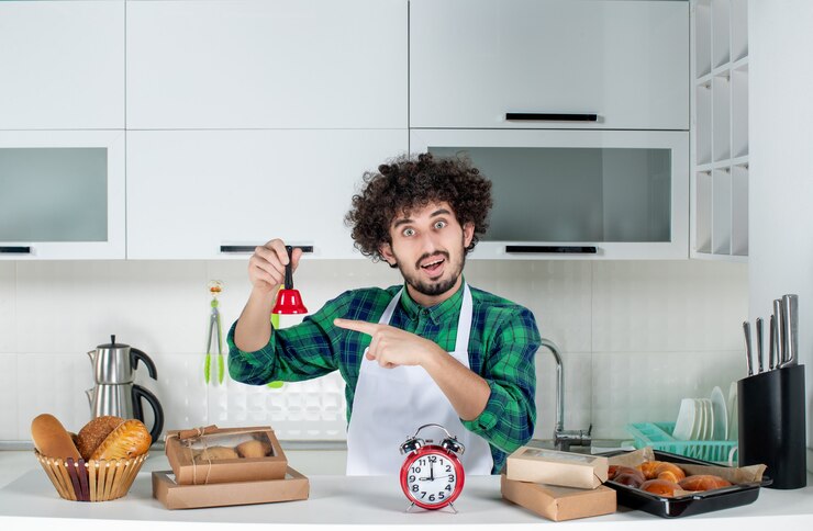 Vista frontal del joven confiado de pie detrás de la mesa varios pasteles y sosteniendo la campana de anillo rojo en la cocina blanca
