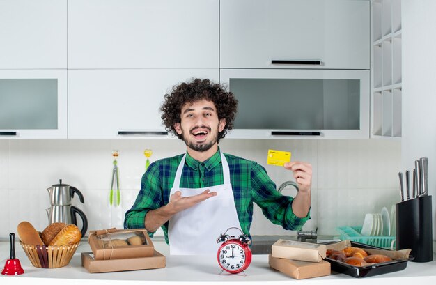 Vista frontal del joven confiado de pie detrás de la mesa varios pasteles y sosteniendo una tarjeta bancaria en la cocina blanca