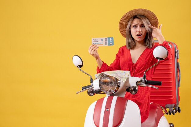 Vista frontal de la joven confundida en vestido rojo sosteniendo boleto poniendo pistola de dedo en su sien en ciclomotor