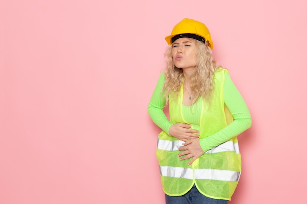 Vista frontal joven constructor en casco de traje de construcción verde que tiene dolor de estómago en la dama de trabajo de construcción de arquitectura espacial rosa