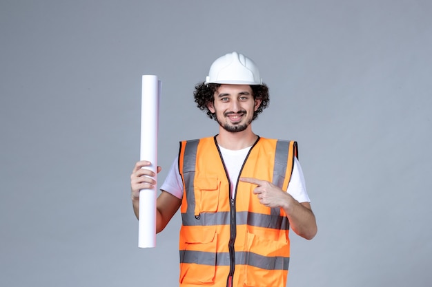 Vista frontal del joven constructor masculino sonriente en chaleco de advertencia con casco de seguridad y mostrando en blanco en la pared gris