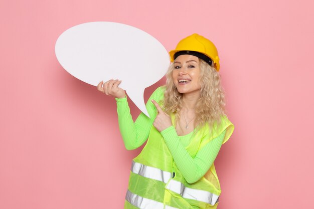 Vista frontal joven constructora en casco de traje de construcción verde sosteniendo un gran cartel blanco con una leve sonrisa en el trabajo de construcción de arquitectura espacial rosa