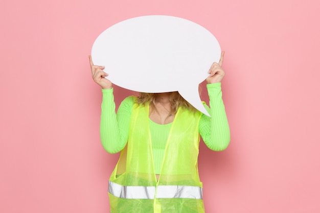 Vista frontal joven constructora en traje de construcción verde casco amarillo con cartel grande blanco en el trabajo de construcción de arquitectura espacial rosa