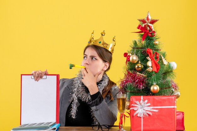 Vista frontal joven con corona sentada en el escritorio sosteniendo documento árbol de Navidad y cóctel de regalos