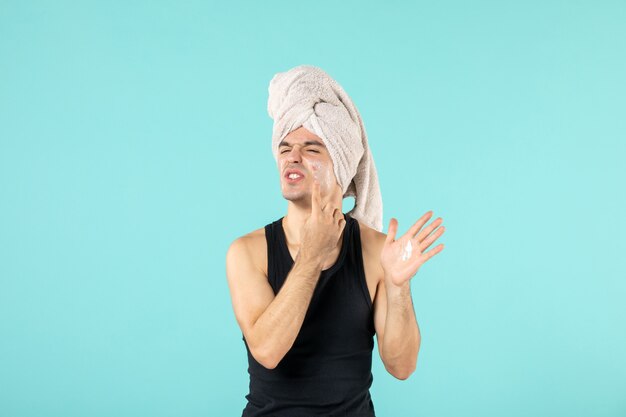 Vista frontal del joven después de la ducha aplicando crema en el rostro en la pared azul