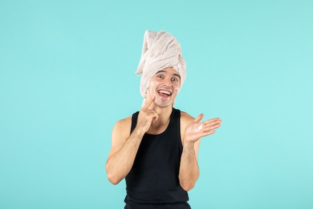 Vista frontal del joven después de la ducha aplicando crema en el rostro en la pared azul