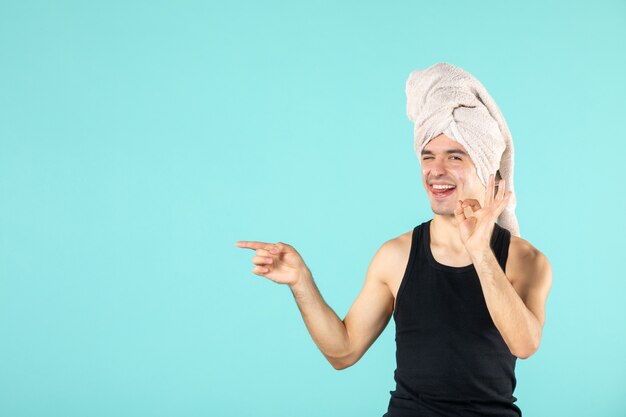 Vista frontal del joven después de la ducha aplicando crema en el rostro en la pared azul