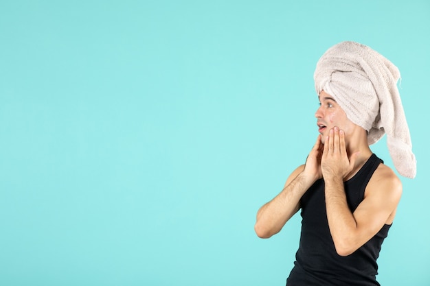 Vista frontal del joven después de la ducha aplicando crema en el rostro en la pared azul