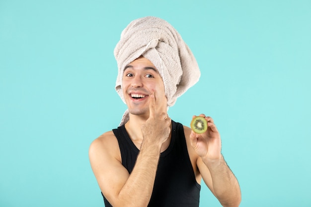 Vista frontal del joven después de la ducha sosteniendo rodajas de kiwi en la pared azul
