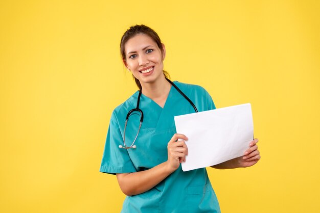 Vista frontal joven doctora en camisa médica con análisis de papel sobre fondo amarillo