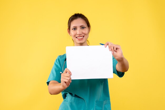 Vista frontal joven doctora en camisa médica con análisis de papel sobre fondo amarillo