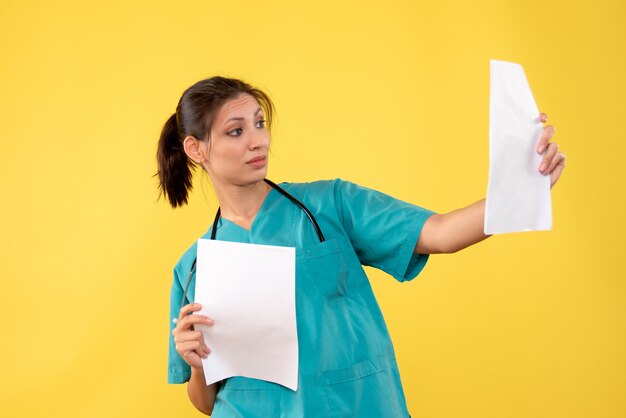 Vista frontal joven doctora en camisa médica con análisis de papel sobre fondo amarillo