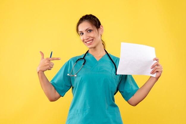 Vista frontal joven doctora en camisa médica con análisis de papel sobre fondo amarillo