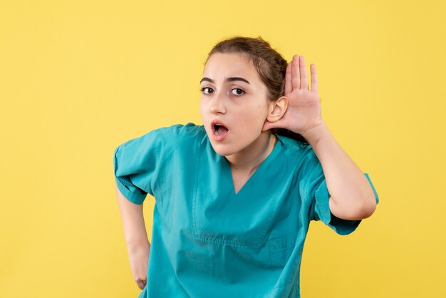 Vista frontal de la joven doctora en camisa médica escuchando en la pared amarilla