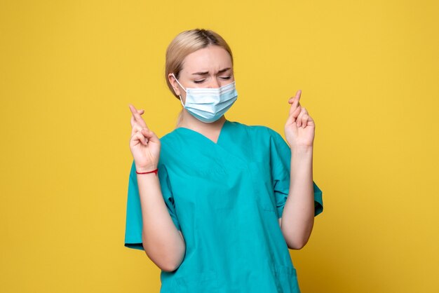 Vista frontal de la joven doctora en camisa médica y máscara estéril cruzando los dedos sobre la pared amarilla