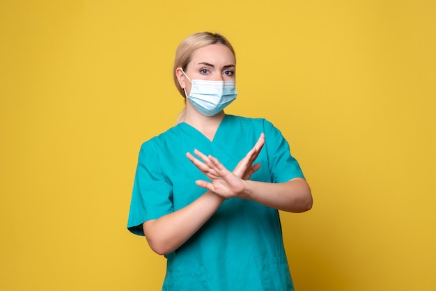 Vista frontal de la joven doctora en camisa médica y máscara estéril en la pared amarilla