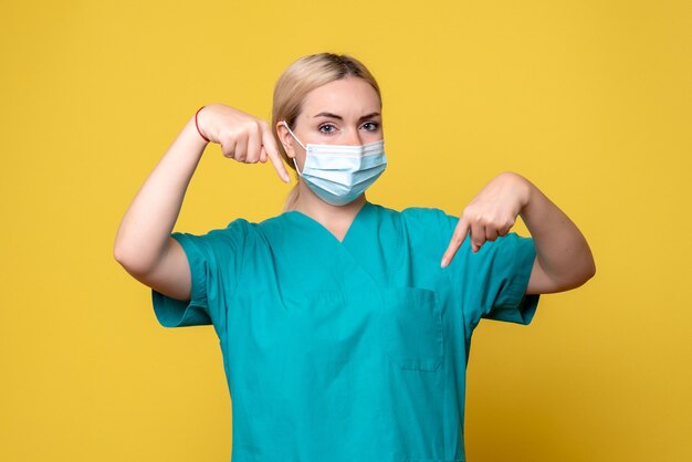 Vista frontal de la joven doctora en camisa médica y máscara estéril en la pared amarilla