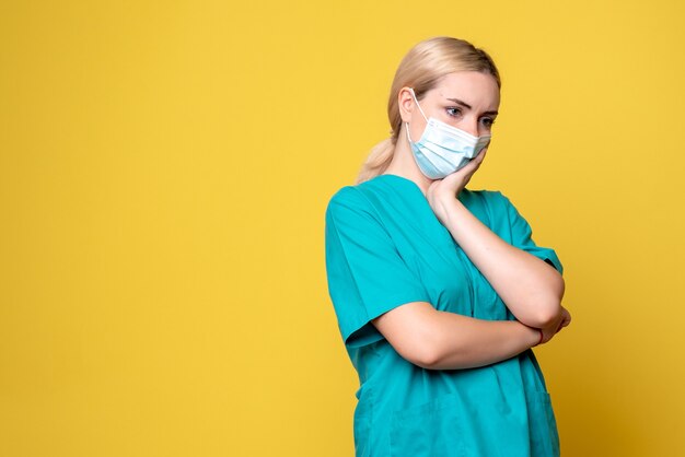 Vista frontal de la joven doctora en camisa médica y máscara en la pared amarilla