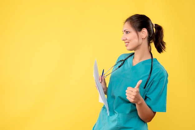 Vista frontal joven doctora en camisa médica con papeles en el escritorio amarillo