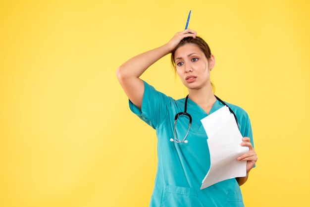 Vista frontal joven doctora en camisa médica con papeles sobre fondo amarillo