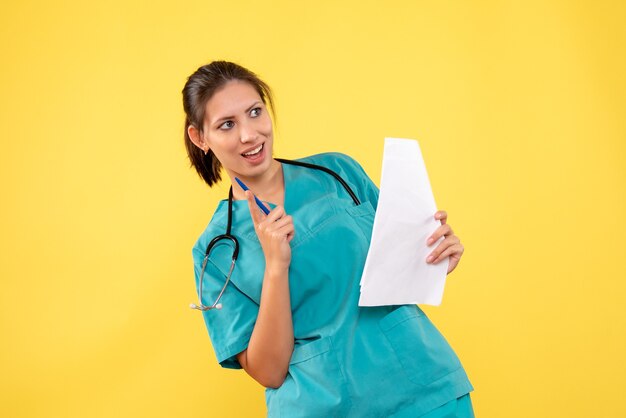 Vista frontal joven doctora en camisa médica con papeles sobre fondo amarillo