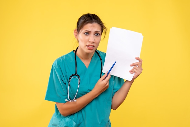 Vista frontal joven doctora en camisa médica con papeles sobre fondo amarillo