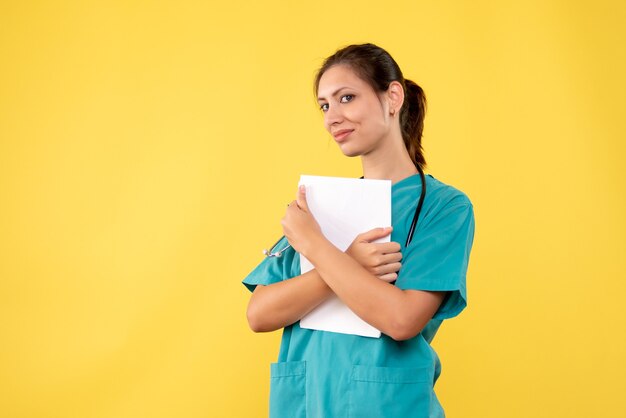 Vista frontal joven doctora en camisa médica con papeles sobre fondo amarillo