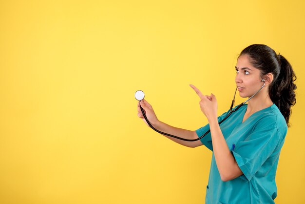 Vista frontal de la joven doctora con estetoscopio apuntando a algo en la pared amarilla
