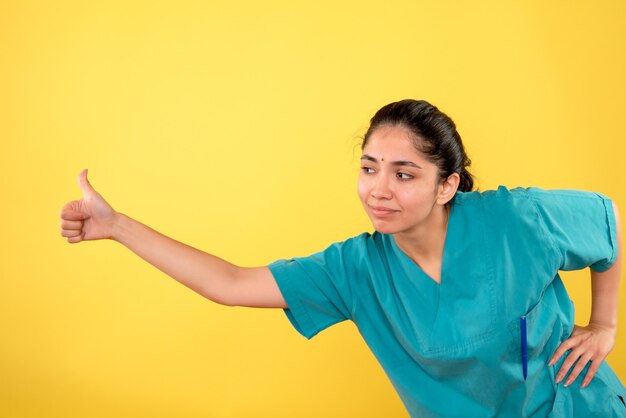 Vista frontal de la joven doctora haciendo pulgar hacia arriba firmar en la pared amarilla