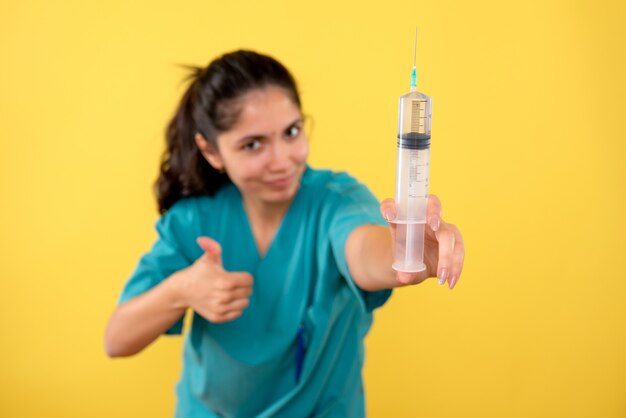 Vista frontal de la joven doctora con jeringa haciendo pulgar hacia arriba firmar en la pared amarilla