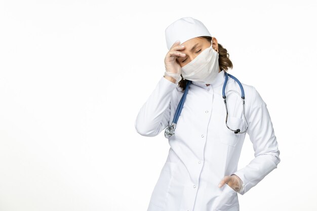 Vista frontal joven doctora con máscara estéril y guantes debido al coronavirus estresado en la superficie blanca