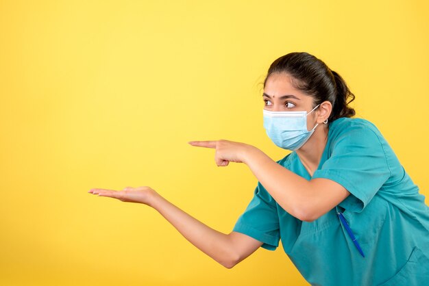 Vista frontal de la joven doctora con máscara médica de pie en la pared amarilla