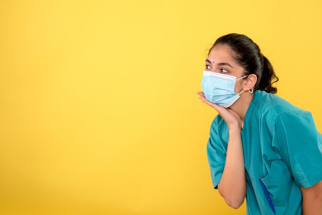 Vista frontal de la joven doctora con máscara médica poniendo la mano en la barbilla en la pared amarilla